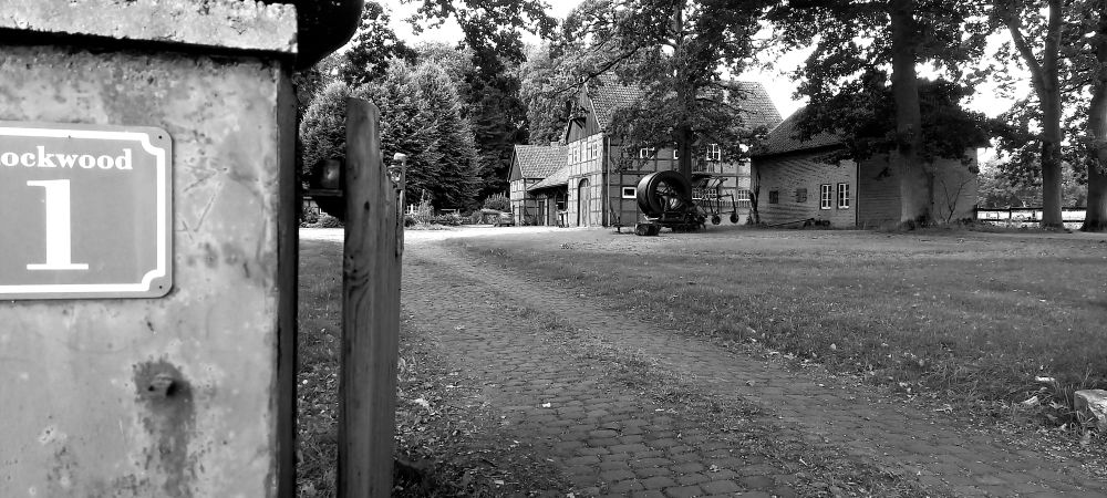 Ein Bauernhof, zu dem ein langer Steinweg führt. Vorne links ist ein Schild zu sehen, das abgeschnitten fotografiert wurde. Lesen kann man "ockwood 1".