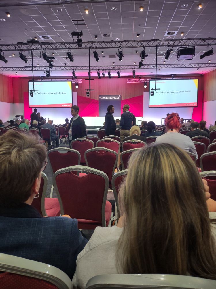 View from a conference seat.  Two adults one with short hair the other with long are in the foreground and with chairs and then screens behind that.  There is a red lighting scheme