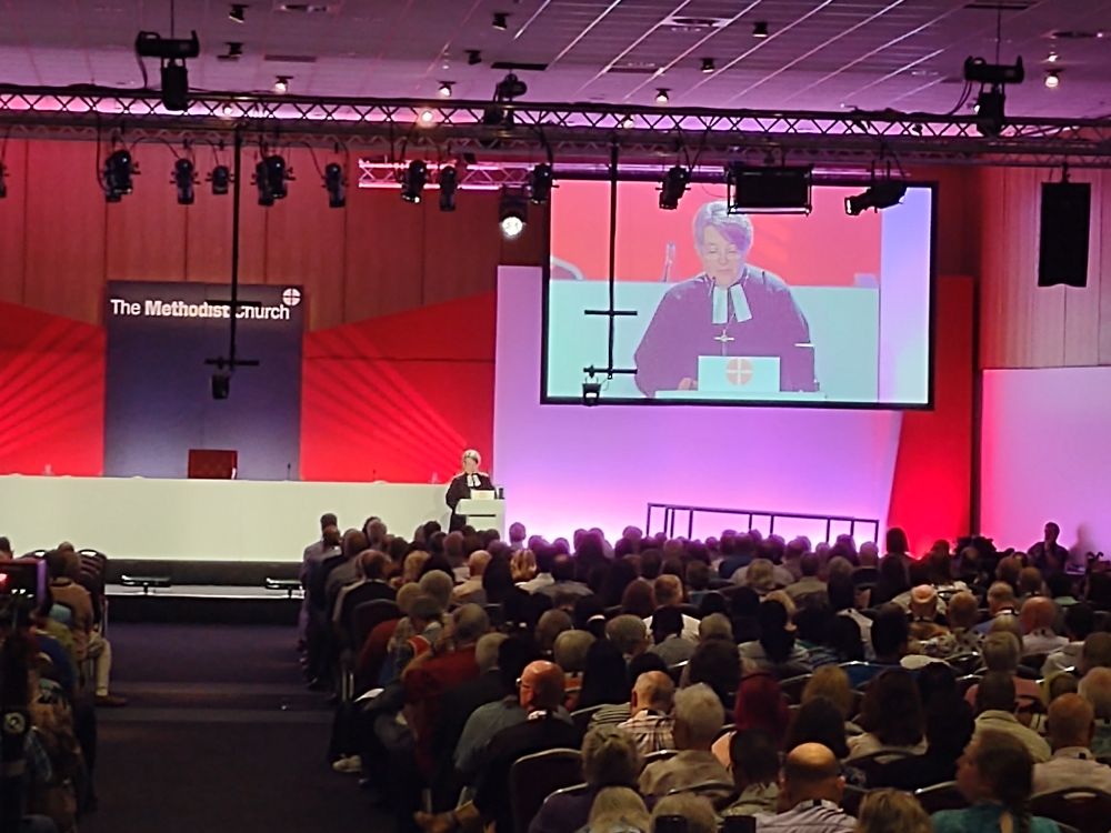 A conference hall from the back and with the presenter on a large screen.  The presenter is a white woman with short hair, wearing a black caasock, preaching gown and white clerical tabs