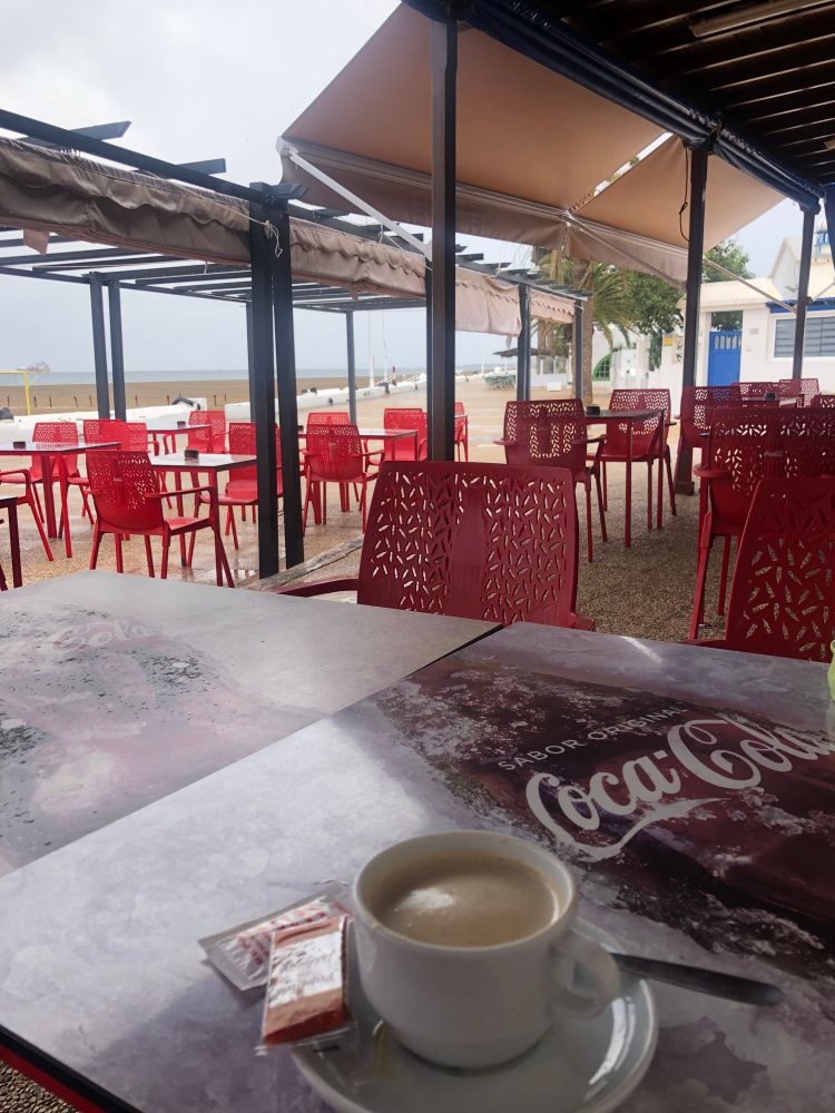 Rustic cafe, empty of people but locals were popping in and out.  It’s next to a sea front promenade with outside seating, but we’re under a canopy.  Red plastic chairs, Cola adverts on the tables, a cup of coffee on our table with a wrapped Speculoos-type biscuit next to it.  If you look closely, there are puddles on the pavement and rain is dripping from the canopies.  You can’t see it, but in the distance a pigeon was having a lovely bath in one of the puddles.