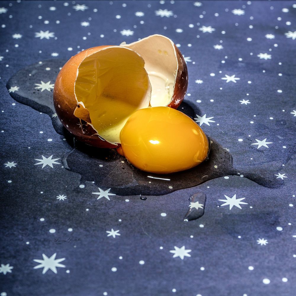 A broken egg with a rich yellow yolk sliding out of one half of the shell. The background is blue with white stars. 
