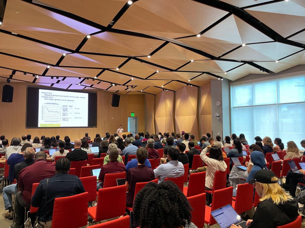 View from the back of the conference room. About 150 people are seated in red chairs while listening to Professor Ken Koedinger. Ken stands at a podium at the front right of the room. A large digital screen projects his powerpoint slide at the front of the room. 