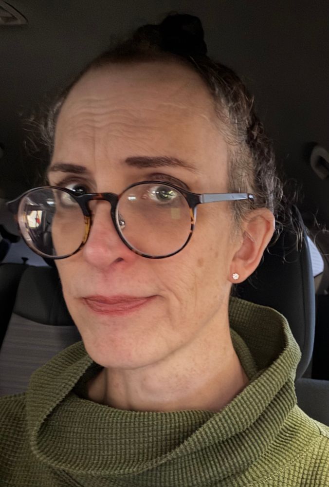 aurora sitting in a parked car, wearing a cowlneck green top, hair tied up, looking tired and cynical