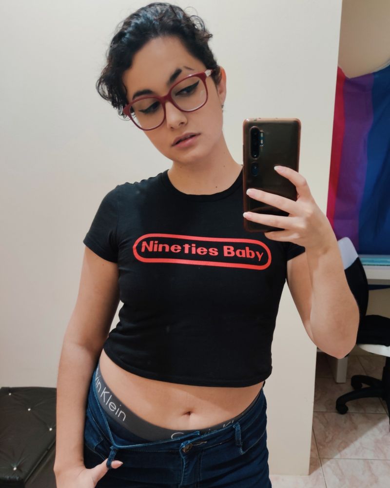 Mirror selfie of Sunny wearing dark jeans getting lightly pulled down to show a Calvin Klein underwear band, a black crop top with "Nineties Baby" written in the Nintendo logo style, red square glasses, and short, black hair.
