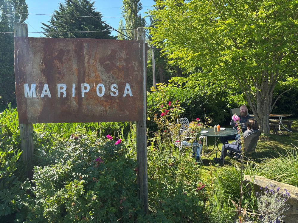 A rustic metal sign for “Mariposa” with an outdoor dining table with many flowers and trees 