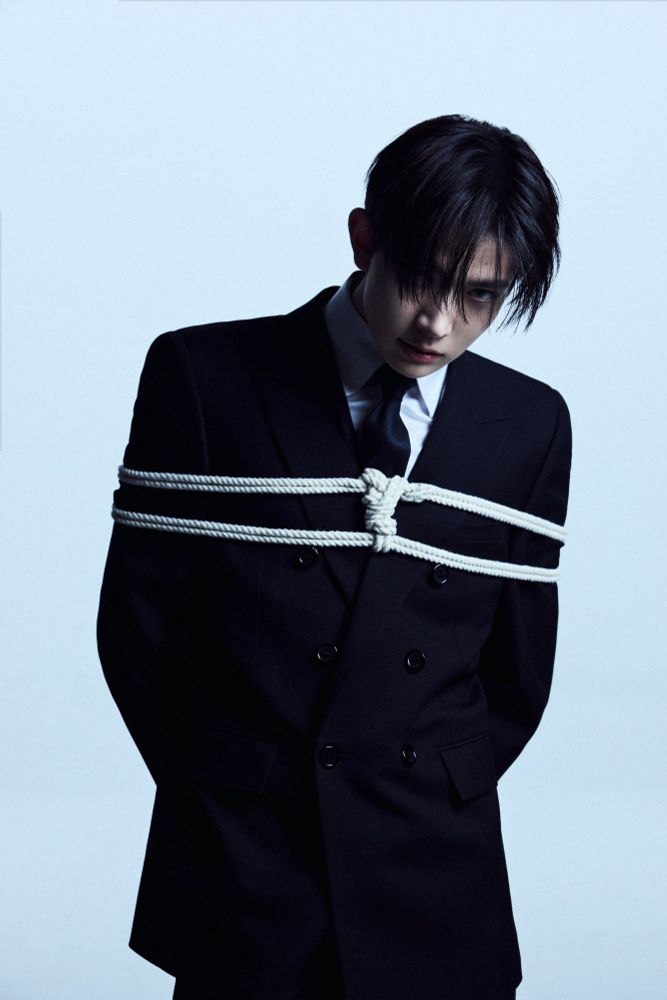lee heeseung (enhypen) olhando a câmera todo sedutor, a franja cai na frente dos olhos. ele está de terno e cordas prendem seus braços de uma forma meio Shibari.