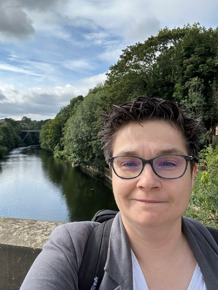 A woman with spiky brown hair, wearing a white t shirt and a grey jacket, with a stunning river backdrop and concrete bridge behind her. 