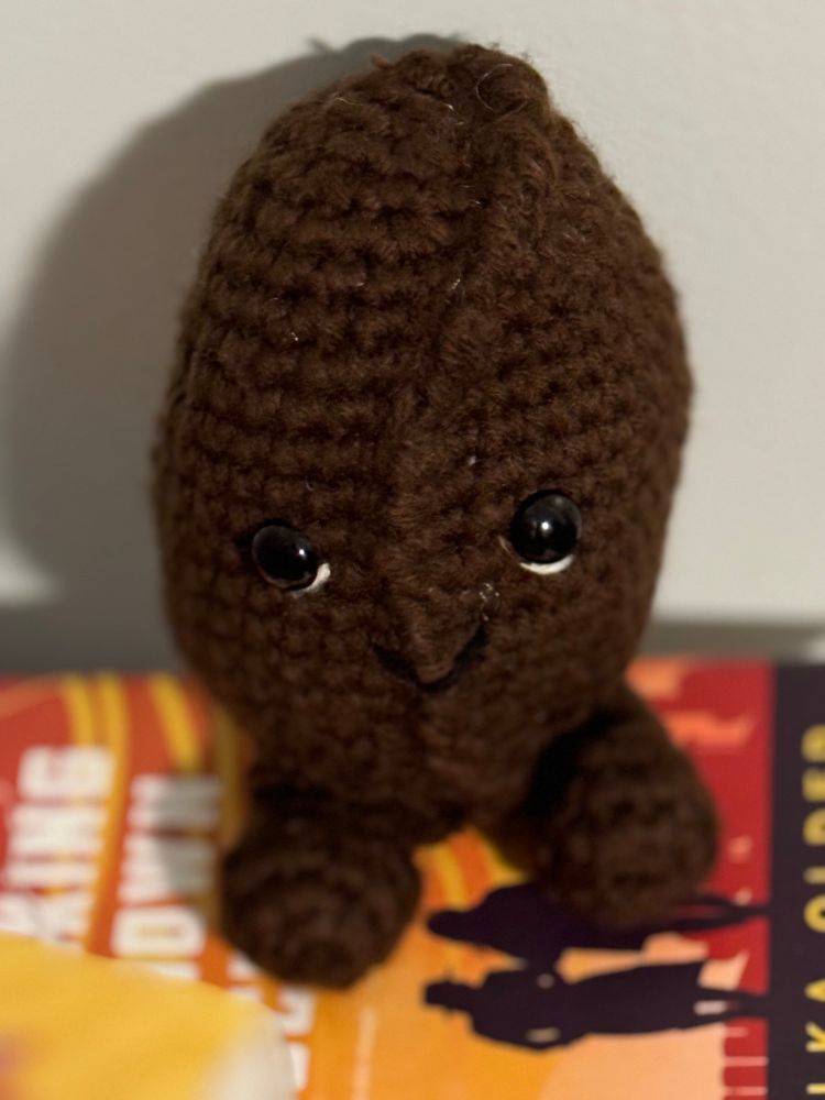 A crocheted coffee bean with a little smile and white lines under his eyes, and little brown feeties.