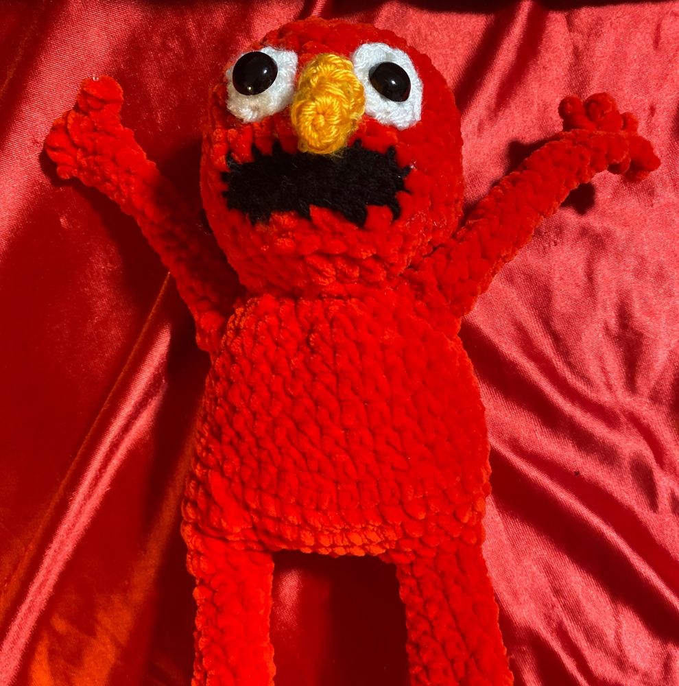 Photo of my crocheted Elmo on a red background, looking real goofy with kind of discombobulated eyes and fingers. Sorry. 