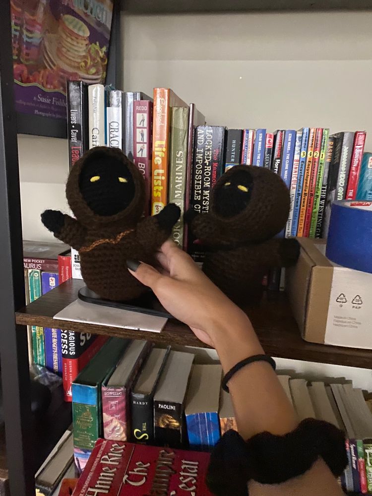 Photo of two crocheted Jawas from Star Wars on a bookshelf. My hand is holding onto the new one, a Jawa with a light brown belt. The Jawa on the right has a black belt. In the background, a a collection of books. 