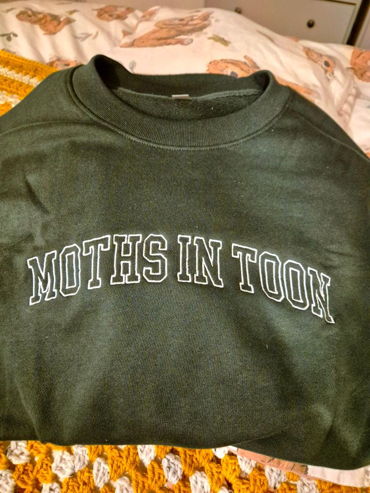 A bottle green sweatshirt laid on a crochet blanket. The sweatshirt is embroidered with the phrase Moths in Toon.