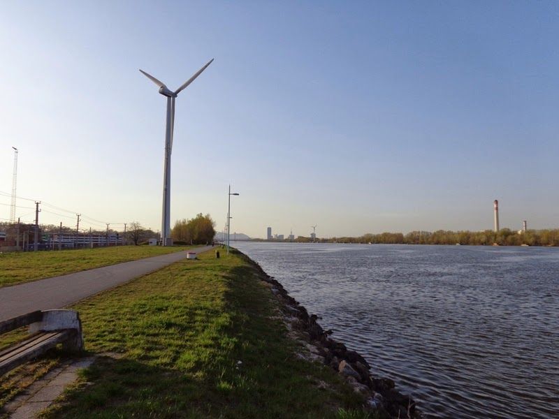 Color photo of the Danube river before sunset, a wind turbine stands tall on the left of the frame as a paved path extends from the bottom left corner towards the center-left of the frame. 