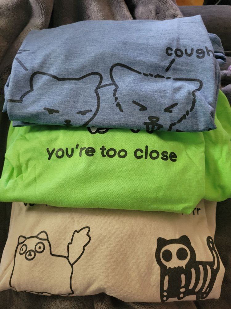 Top to bottom: -a heather cornflower tee with kitties -a neon green tee with a kitty saying you're too close -A sand colored tee with some kitties dressed up for Halloween (2 shown a ghost and a skeleton)