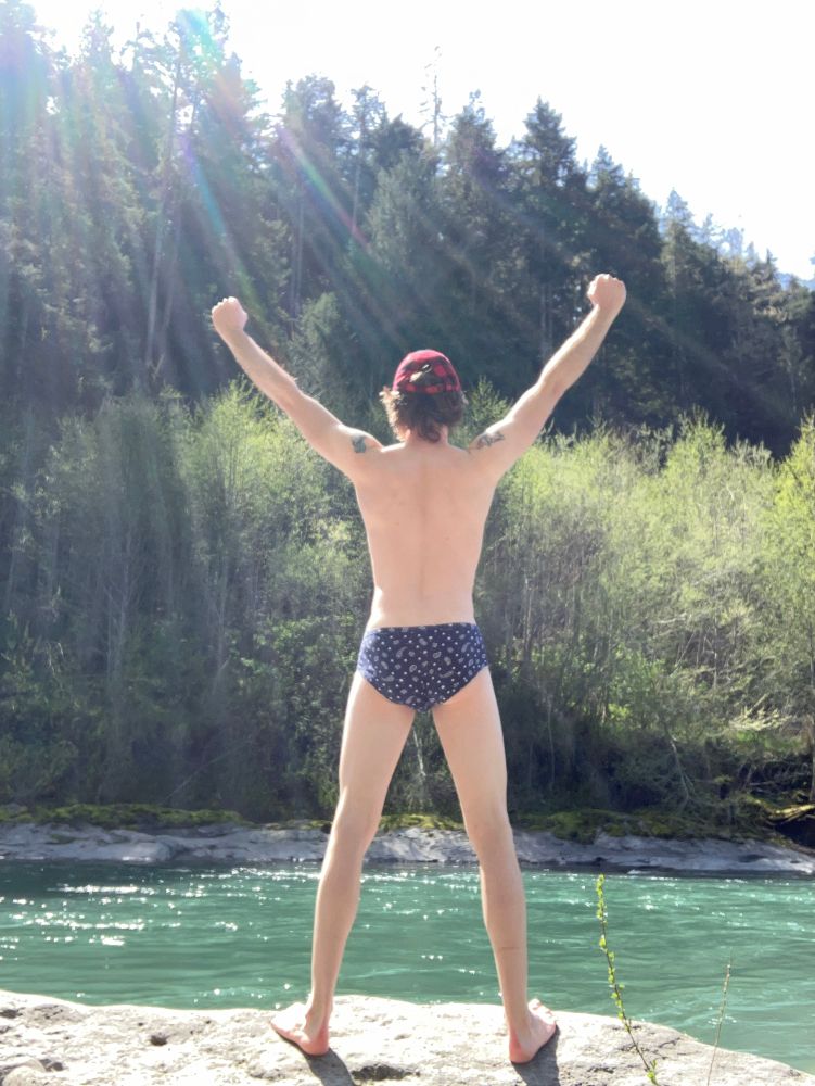 Person standing by a river with arms raised above their head facing away from the camera 