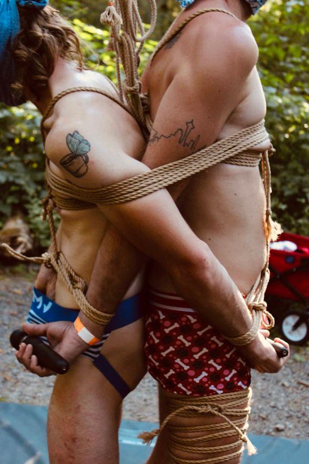 Two masculine bodied people tied together with ropes outside in the forest while holding vibrating wands against each other