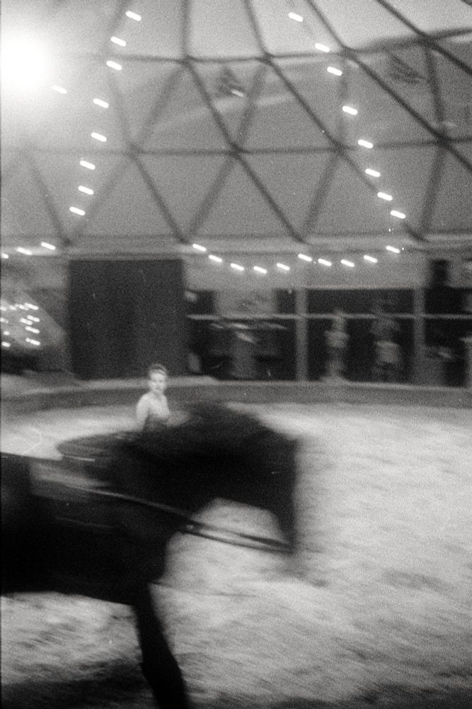 Photographie en noir et blanc d'un cheval de voltige en mouvement, flou. Une écuyère est au milieu de la piste. 
photographie argentique en noir et blanc, Kentmere400