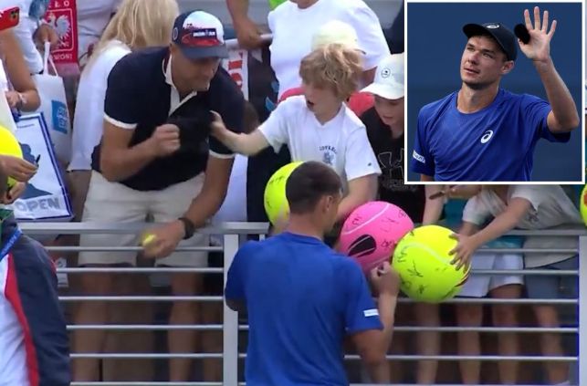 Tennis player Kamil Majchrzak signing autographs after winning a match. Clad in blue kit from Ascics. Has just giving his hat to a kid in the stands in a white t-shirt. A douchecanoe paving stone CEO has snatched the hat away from the kid and is about to hand it to his wife to put in her bag. 