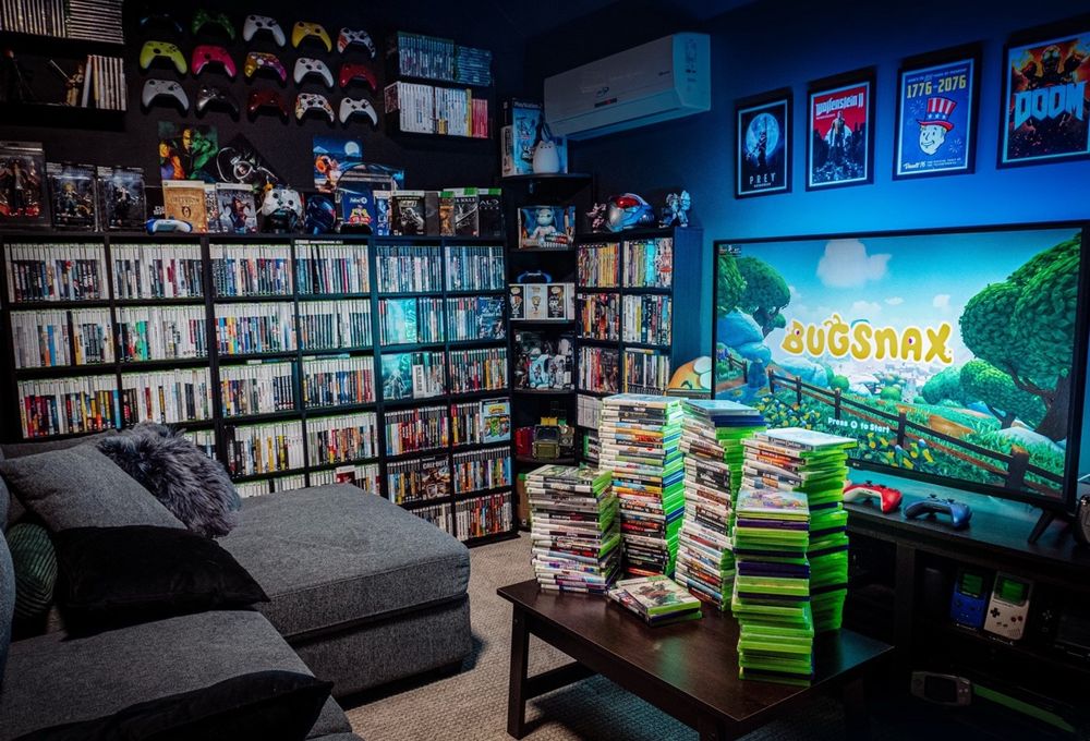 Mo’s game room with various physical video games for the Xbox, Xbox 360, PlayStation 1, 2, & 4, GameCube, and various Xbox One controllers. Bugsnax is on the TV