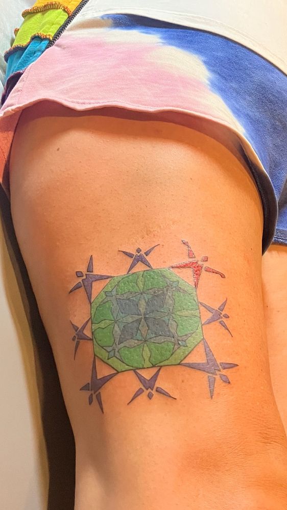 A green, blue and purple geometric and quilt like octagon design with blue people on the edge, and one bright orange/red flapping person in the top right corner. This is a tattoo on the leg , just above the knee and on the side of a tanned colored white person’s leg. 
