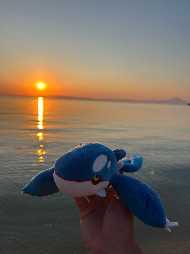 A hand holding a Kyogre plushie in front of a backdrop of the sun rising above a gentle sea