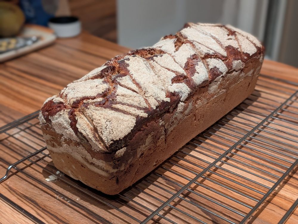 
A rectangular loaf of bread with a light brown, slightly cracked crust sits on a black metal cooling rack, which rests on a wooden countertop. The bread looks rustic and homemade.
In the background, there is a cream-colored wall and a light-colored wooden floor.
