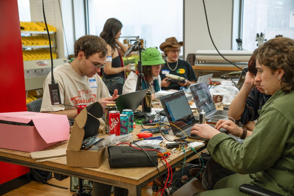 Hackers in the fabrication studio, with the nearest hacker wearing a green jumpsuit surrounded by vapes hooked up to multimeters and an Arduino