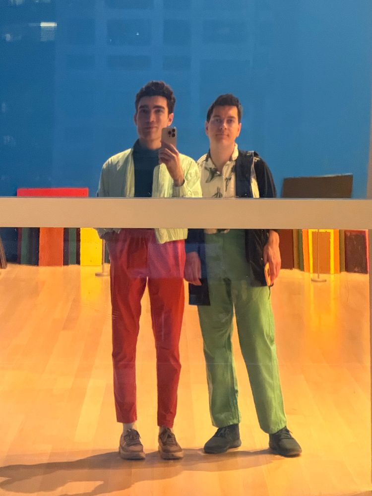 Portrait of Lachlan and Matthew Stanciu in vibrantly colorful outfits in a mirror, which is visually obscuring their waistlines, with colorful blocks on a wood floor and a bright blue background