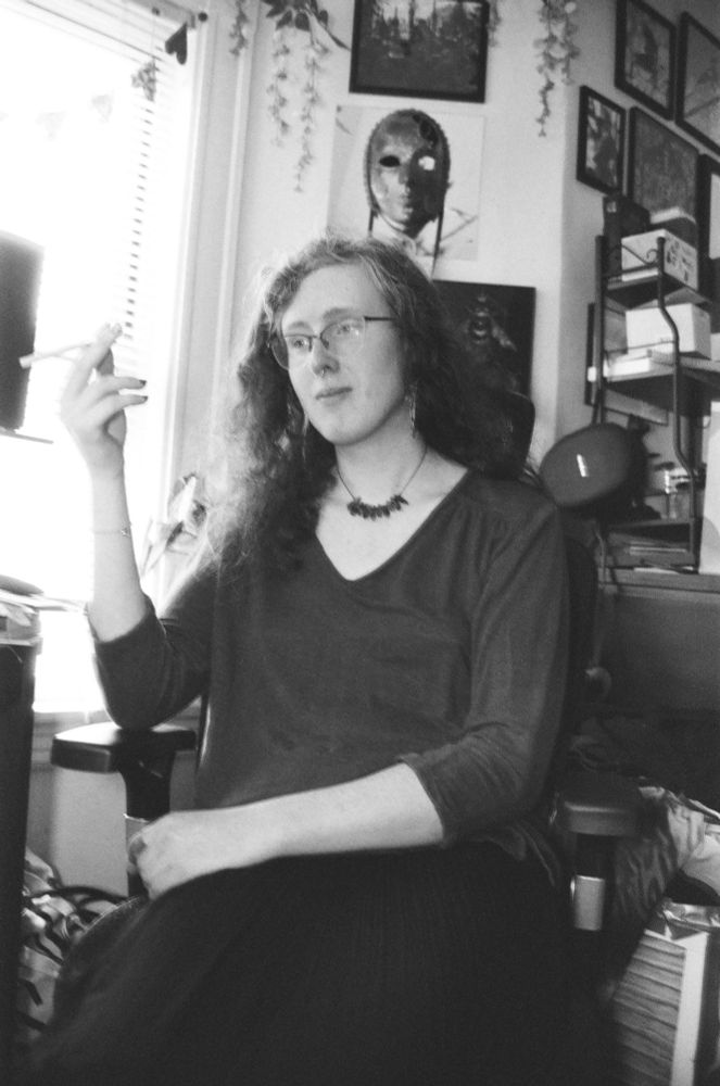 A black-and-white orthochromatic picture of a woman with curly hair wearing a necklace and v-neck blouse with a skirt. She's holding a cigarette aloft but it is unlit