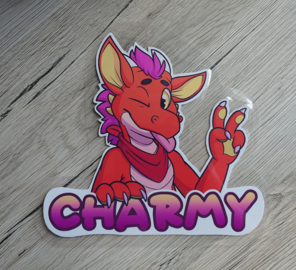 Photo of a con badge showing an orange dragon with pink hair behind the name "Charmy". He is winking at the viewer, showing his tongue and doing the peace sign with his hand.