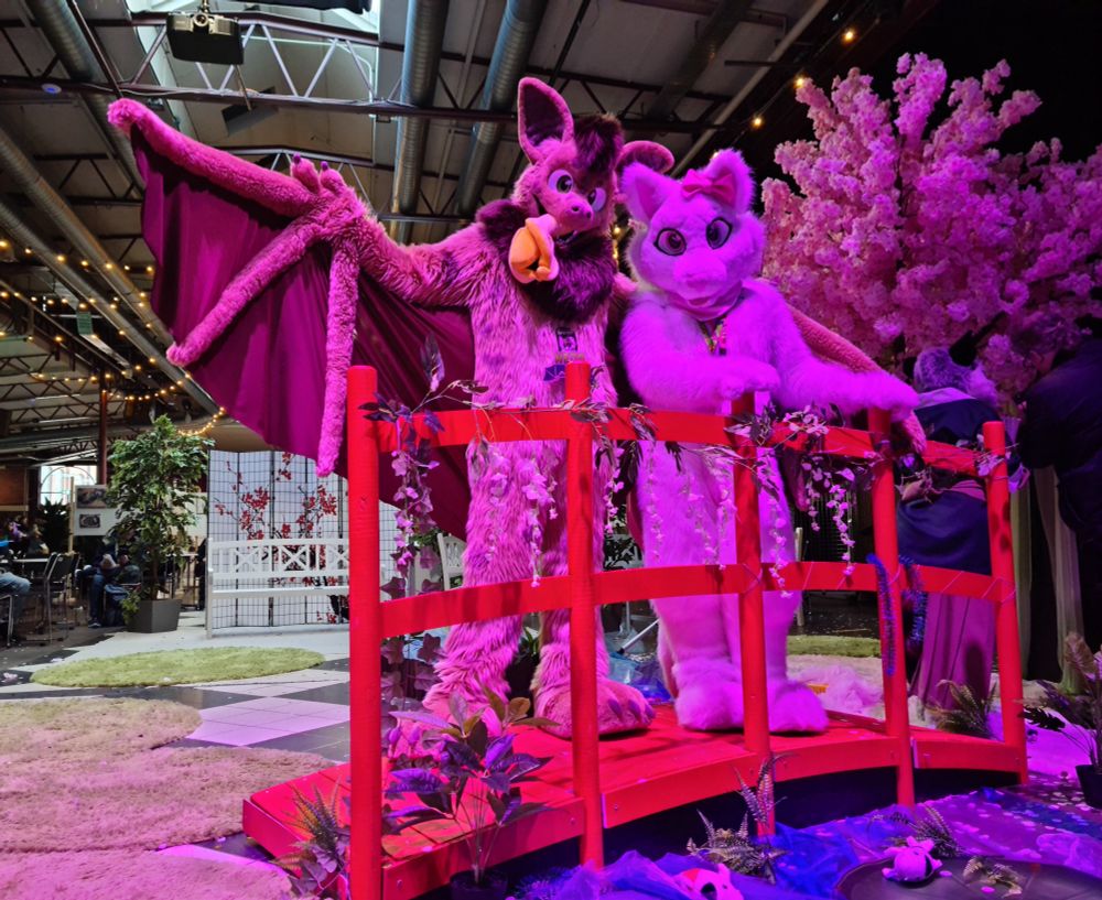 Photo of Neop the bat and Akemi the white cat (fursuits) standing on a red bridge over some water made from cloth with plush fishes. There is a blossoming cherry tree in the near background. Pink light makes them look very pink. The further background shows that they are inside an event hall.