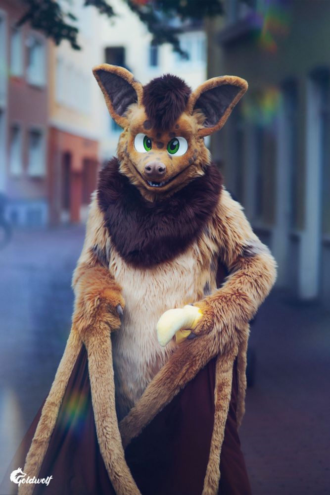 Photo of Neop the bat (fursuit), looking at the viewer and holding a plush banana in one of his wings. The blurry background shows a paved street and some buildings of a German town. The photo generally looks rather soft and dreamy.
