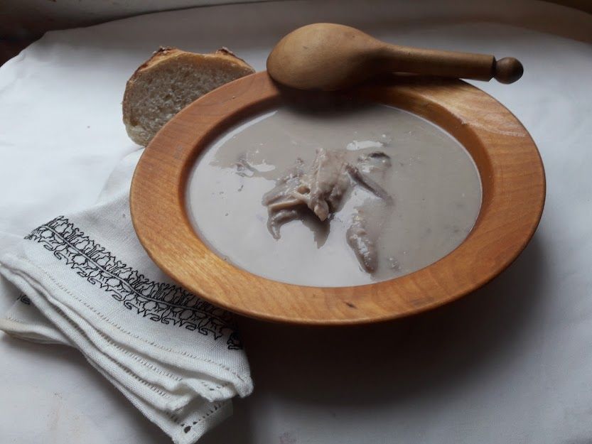 Photo of a plate of soup, arranged for photography. The light beige, opaque soup with pieces of chicken visible in it is served in a wooden plate. A wooden spoon rests on its rim, a cut bread roll and an embroidered napkin surround it. White linen serves as the background and we can just see, in the top left corner, that it is in fact a folded sheet and the whole thing is advertising. We actually ate it with rice, from colourful porcelain plates. 