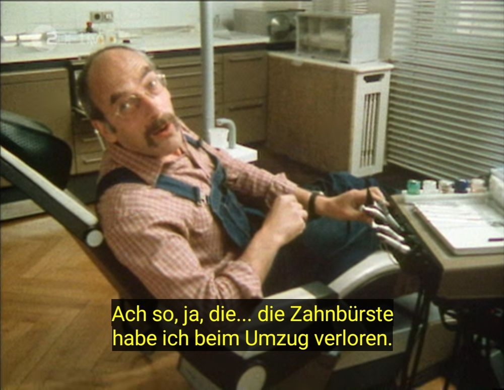 Einzelbild. Peter Lustig sitzt im Behandlungsstuhl beim Zahnarzt und dreht sich leicht zur Kamera. Im Untertitel steht: "Ach so, ja, die... die Zahnbürste habe ich beim Umzug verloren."