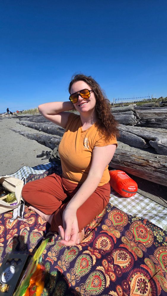 Heather in orange pants and a yellow shirt, sitting on a mandala tapestry at the beach.