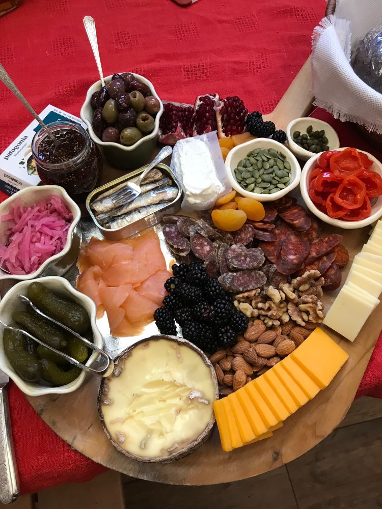 A wooden board on a red tablecloth covered with cheeses, sausages, cured fish, fruit, nuts, and pickles. 