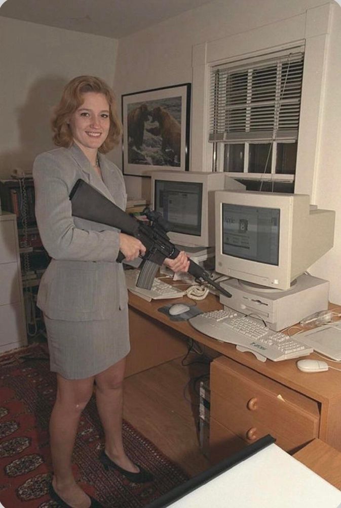 A redheaded white woman evidently cosplaying as Dana Scully smiles and points a gun at two early-2000s looking computers. I do not know who she is, who took this photograph, or how it came to be on my phone