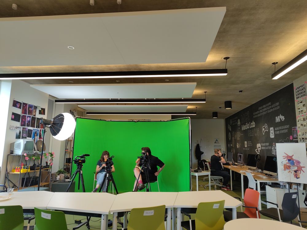 Two figures in front of a green screen in the CMPS media lab. A third lurks in the background 