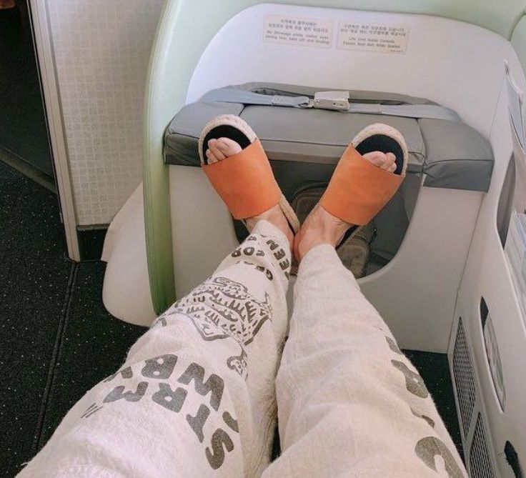 photo hobi took of his feet in a plane. he's got grey sweats on with lettering, and is wearing orange slides. he's got a shallow lil first position going on
