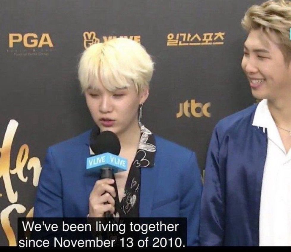 A picture of blonde yoongi talking into a mic saying “We’ve been living together since November 13 of 2010” while Namjoon is off to the side, dimples out, looking fond as fuck