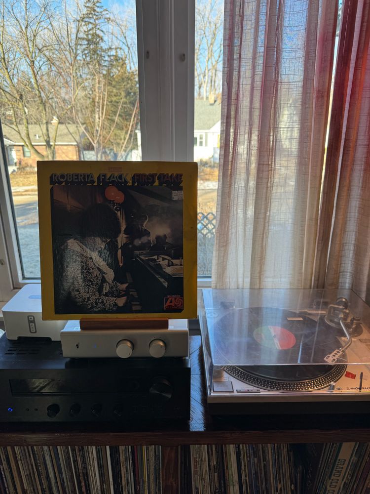 A low record cabinet, you can see the tops of several LPs. A silver turntable sits on top of the cabinet, next to a black stereo receiver and silver phono preamp. An album cover is propped on a stand on top of the preamp: “First Take”, by Roberta Flack. The cover shows Ms Flack, a young Black woman, seated at a baby grand piano in a smoky, wood-paneled room; she wears her hair in an Afro and has on an _excellent_ floral print top - maybe a jacket? And a white lacy scarf. Behind her, partially obscured by smoke and backlit, are two Black men. One is standing and playing an upright bass, and wearing a suit and tie; the other is seated. He’s mostly blocked by the piano but he’s probably at a small drum kit. The whole vibe is just cool as hell.