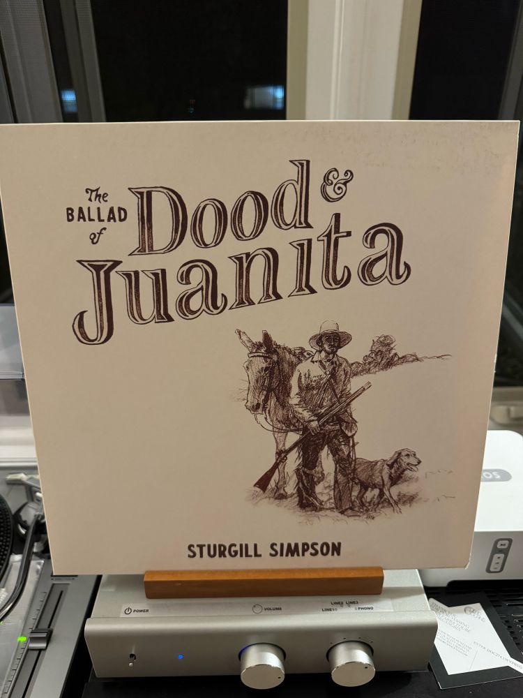 The record album sleeve for Sturgill Simpson’s “The Ballad of Dood and Juanita”. It’s a cream color; the title is done in old-timey Western-style type. In the bottom right is a monochrome sketch of a man holding a rifle, standing in front of a large mule. A dog is at his side.