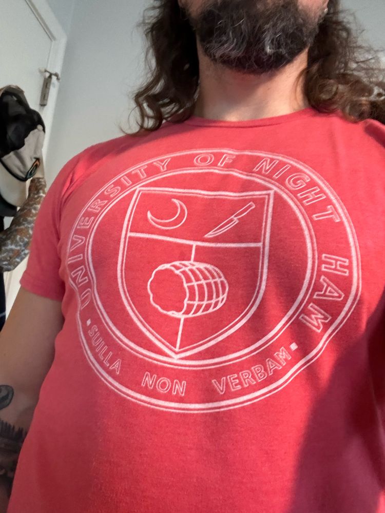 A bearded man wearing a “University of Night Ham” tshirt. You can’t see his face. The shirt has a coat of arms with a crescent moon, a knife, and a ham, and bears the motto “SUILLA NON VERBAM”