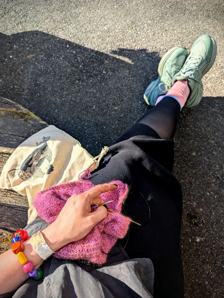 View from above of legs and feet with a hand holding knitting needles and a knitting project