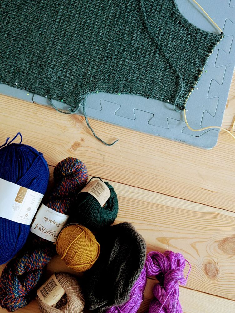 A green knitted piece pinned on a blocking mat and a bunch of yarn laying on a wooden floor.