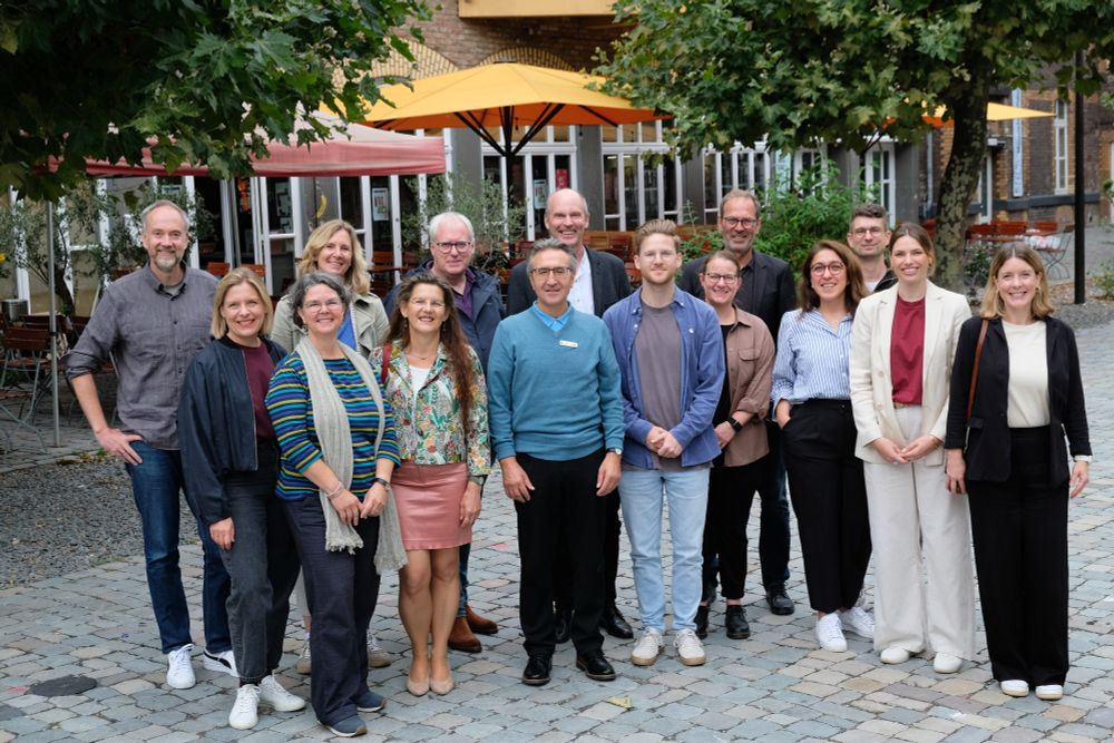 15 Personen stehen auf einem gepflasterten Platz. Im Hintergrund sind Bäume, aufgespannte Sonnenschirme und eine Hausfassade mit großen Fenstern zu sehen. Die Menschen haben sich für ein Gruppenbild aufgestellt und lächeln in die Kamera.