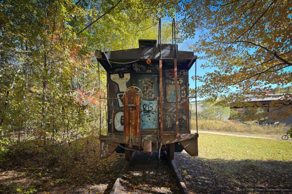 The rear end of a rusted train car sits idle on a short stretch of track in a wooded area, its surface covered in colorful graffiti. Autumn trees surround the scene, and a dirt path runs parallel to the tracks. In the background, a cyclist rides through the forest, adding a touch of motion to the otherwise quiet, sunlit setting.