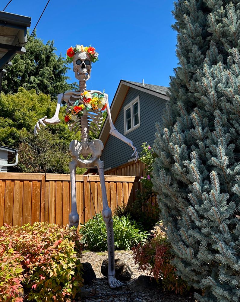 Giant white skeleton standing outdoors wearing a crown and necklace of colorful plastic flowers
