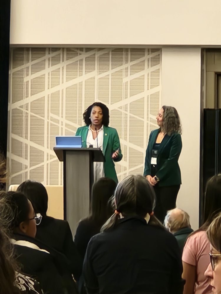 Two women standing on a stage, one behind a podium and the 2nd standing at her left. Audience onlookers are visible in the foreground of the picture.