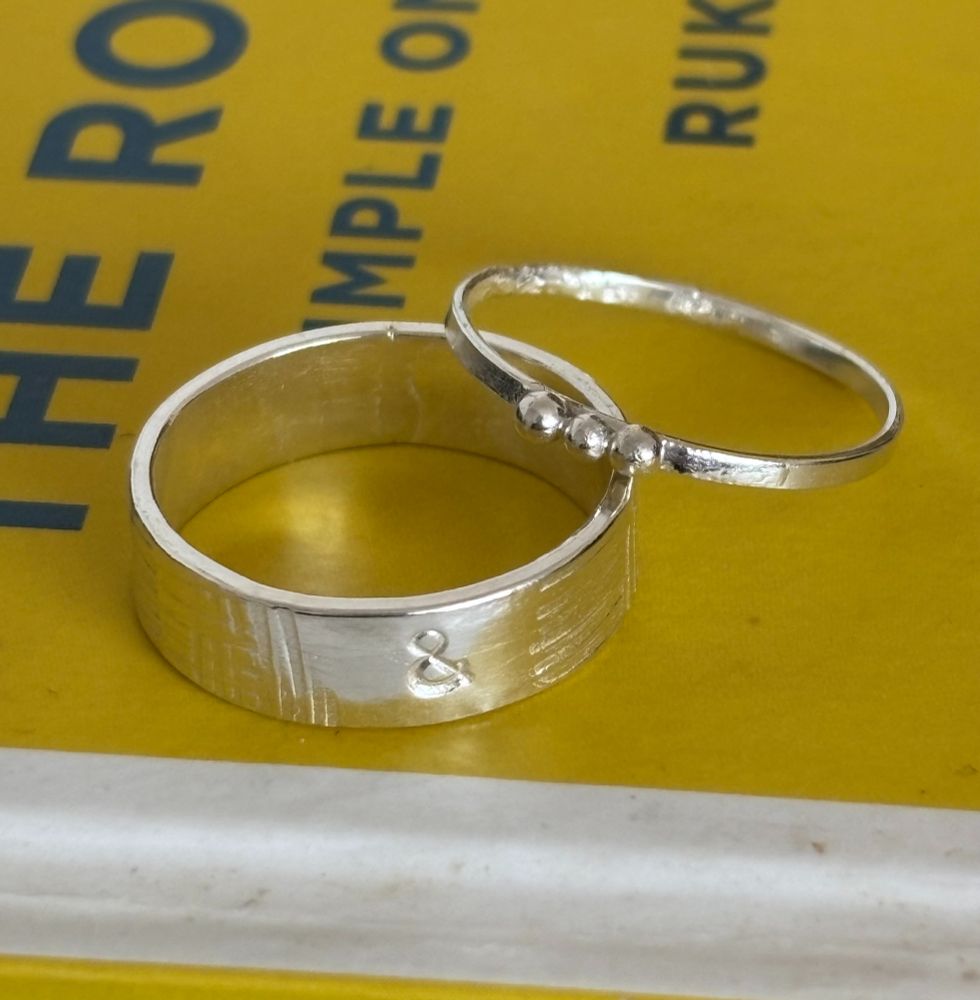 Two silver rings on top of a yellow book. One has a wide band and is stamped with an ampersand, the other is thin with three small silver balls.