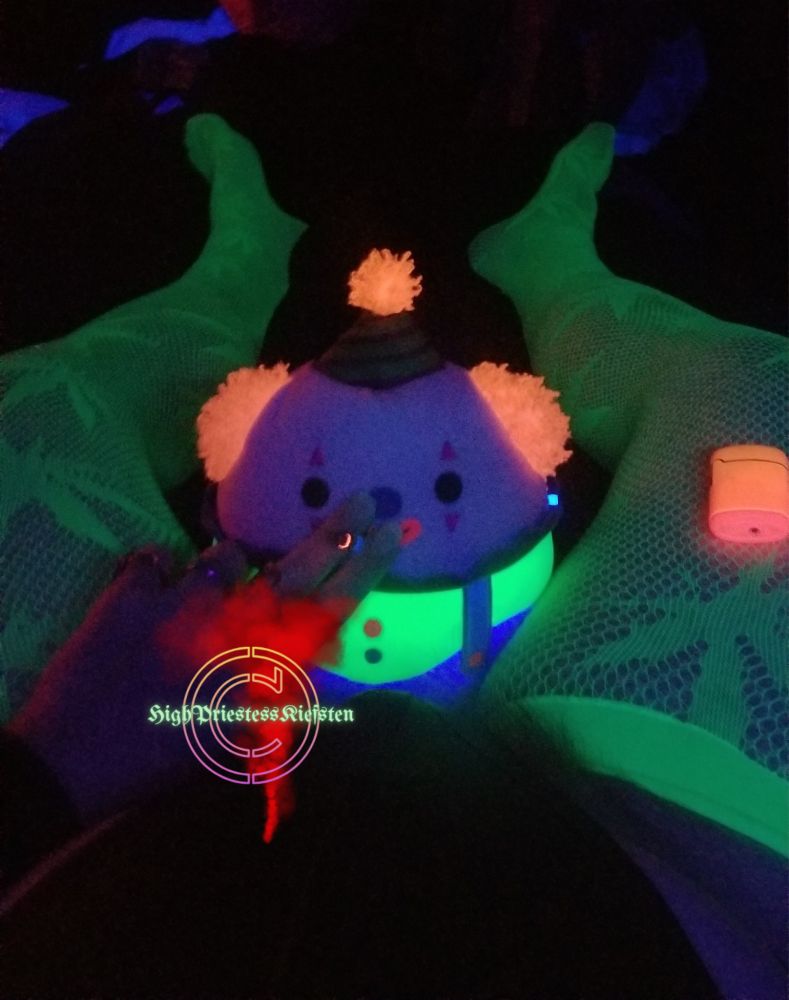 Umberto the Clown smoking a joint with High Priestess Kiefsten, Trans Emo Domme Terror, seated between her thighs. She's wearing a bodysuit & pot leaf thigh highs.  They're glowing together under the blacklight. 